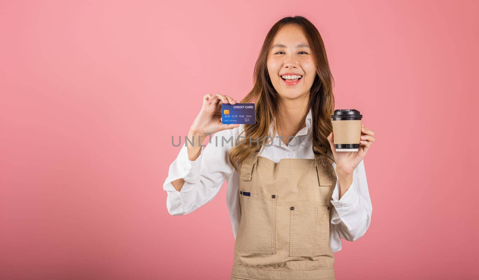 Portrait Asian happy young woman barista bar tender coffee maker holding coffee tea hot cup and credit card studio shot isolated on pink background, female smiling hold takeaways cup for payment