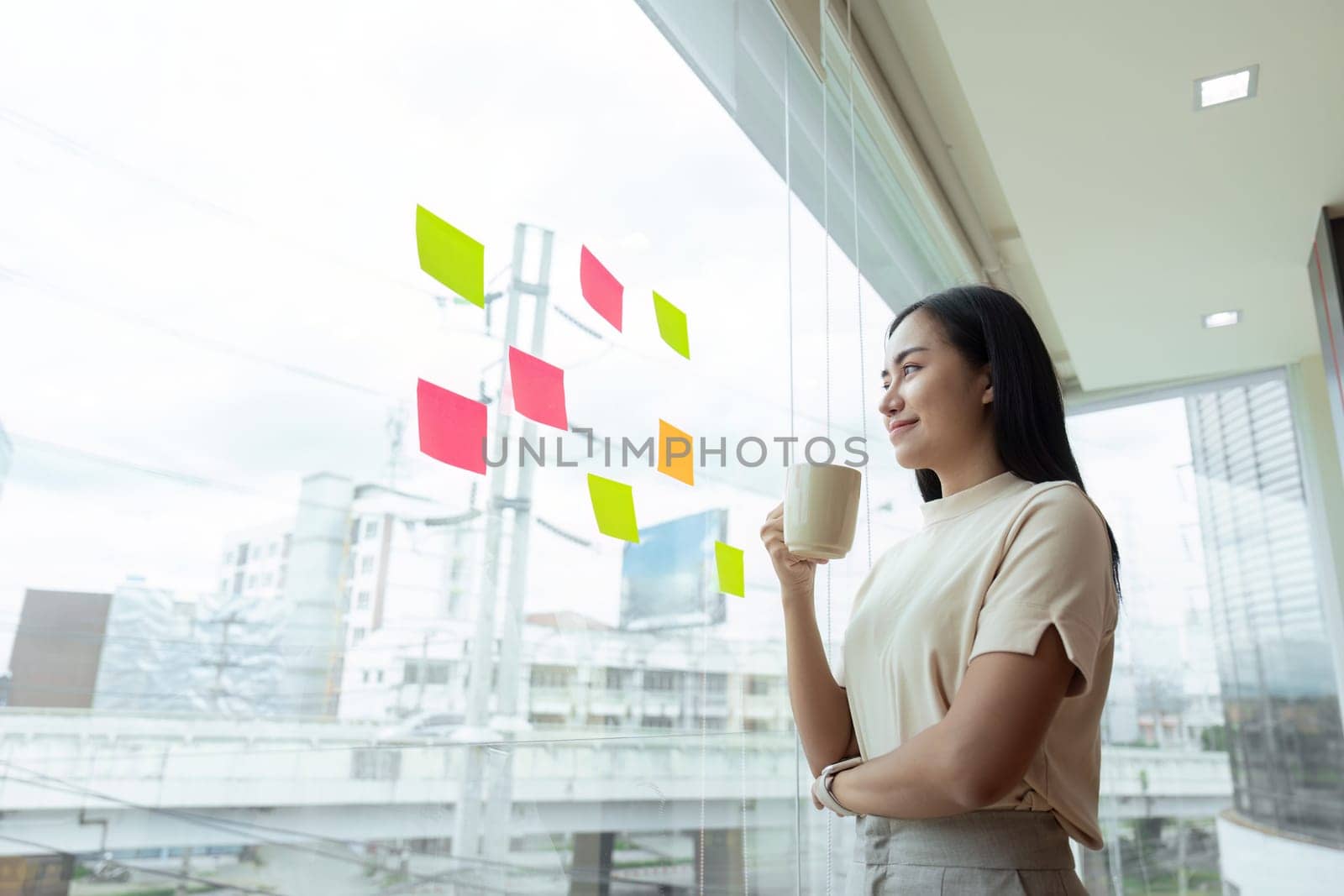Businesswoman brainstorming ideas while looking at sticky notes on a window in a modern office setting, focusing on planning and creativity