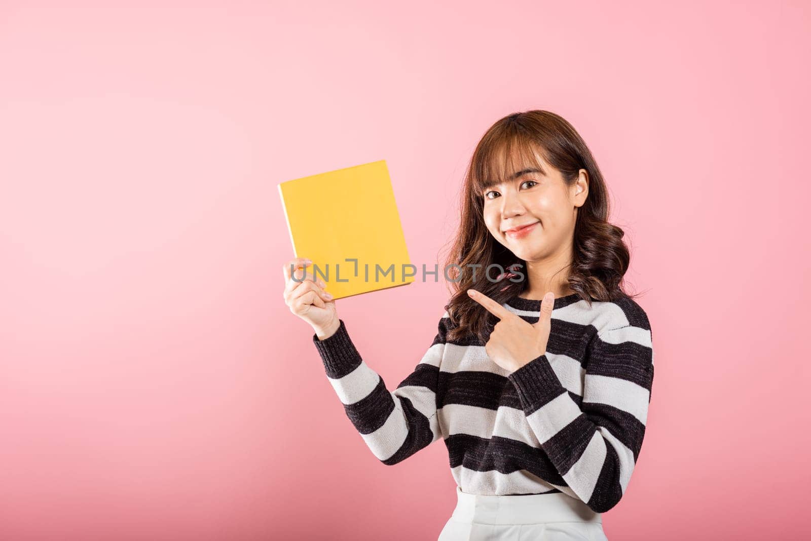 Portrait of happy Asian beautiful young woman confident smiling holding yellow book and pointing finger to the book, studio shot isolated on pink background, education concept
