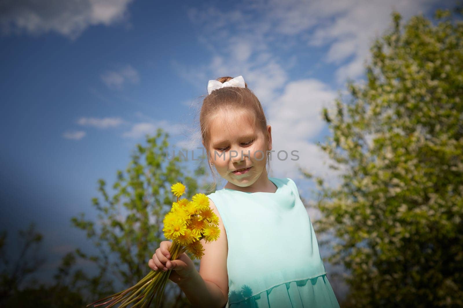 Little girl with a bouquet of yellow dandelion flowers and green trees with a blue sky in the background. Portrait of a happy Baby on a sunny summer day in nature