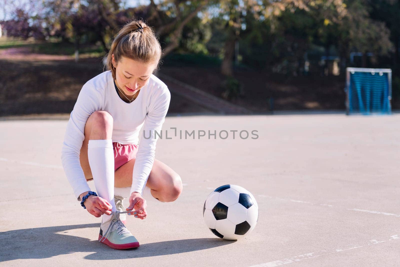 young caucasian woman tying her sneakers next to a soccer ball to play in a urban football court, concept of sport and active lifestyle, copy space for text