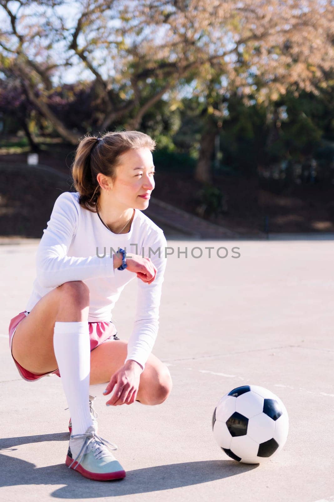 young caucasian woman squatting next to a soccer ball ready to play in a urban football court, concept of sport and active lifestyle