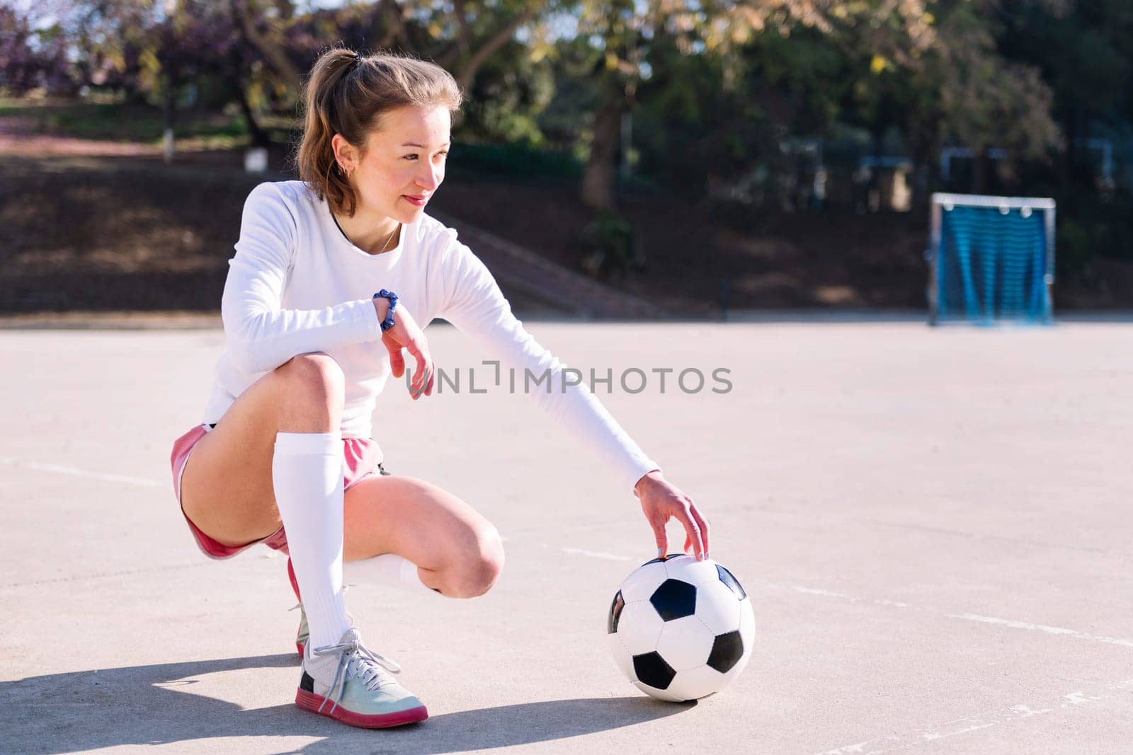 young caucasian woman squatting next to a soccer ball ready to play in a urban football court, concept of sport and active lifestyle