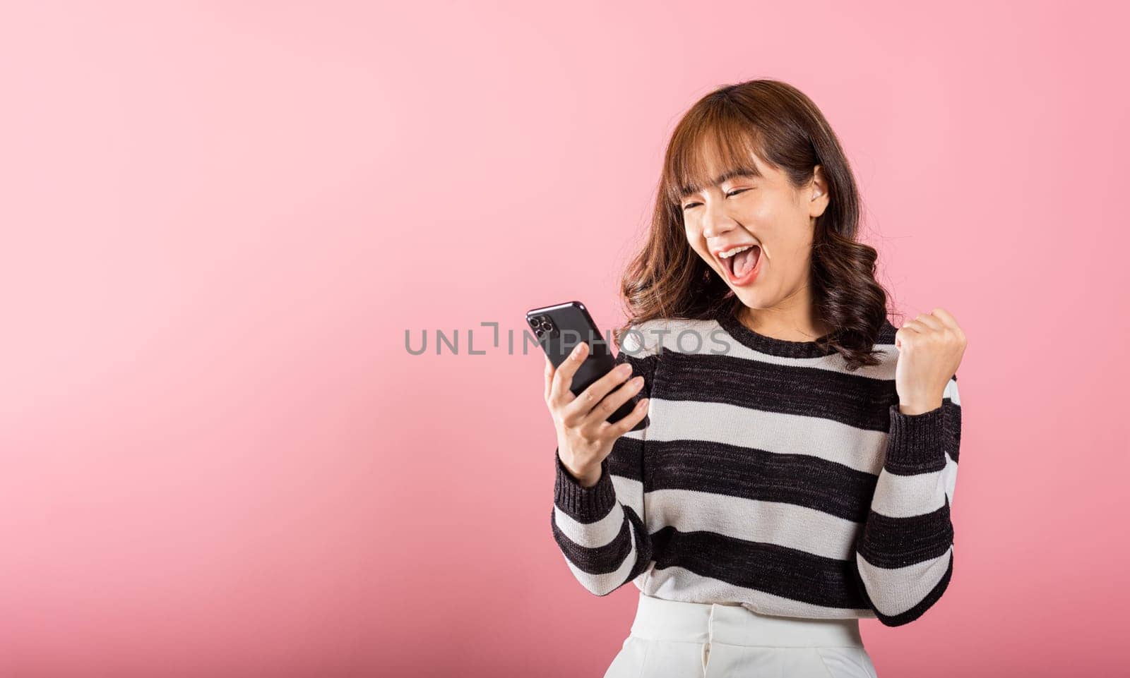 Happy Asian portrait beautiful cute young woman teen smiling excited using mobile phone say yes Studio shot isolated on pink background, Thai female surprised make winner gesture on smartphone