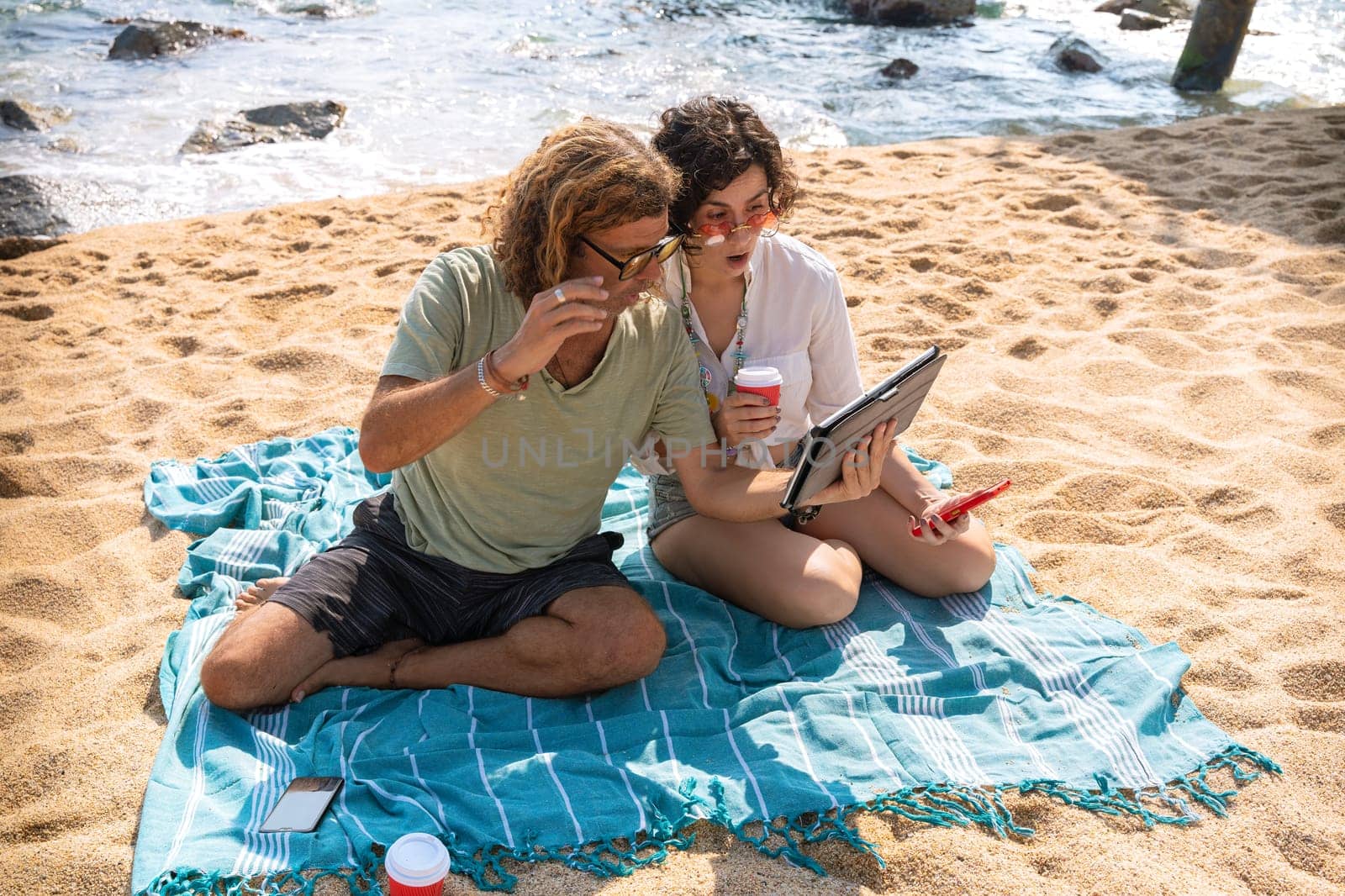 Middle-aged man and woman sitting on the beach with surprised face, browsing tablet apps. Concept:Holidays y Technology