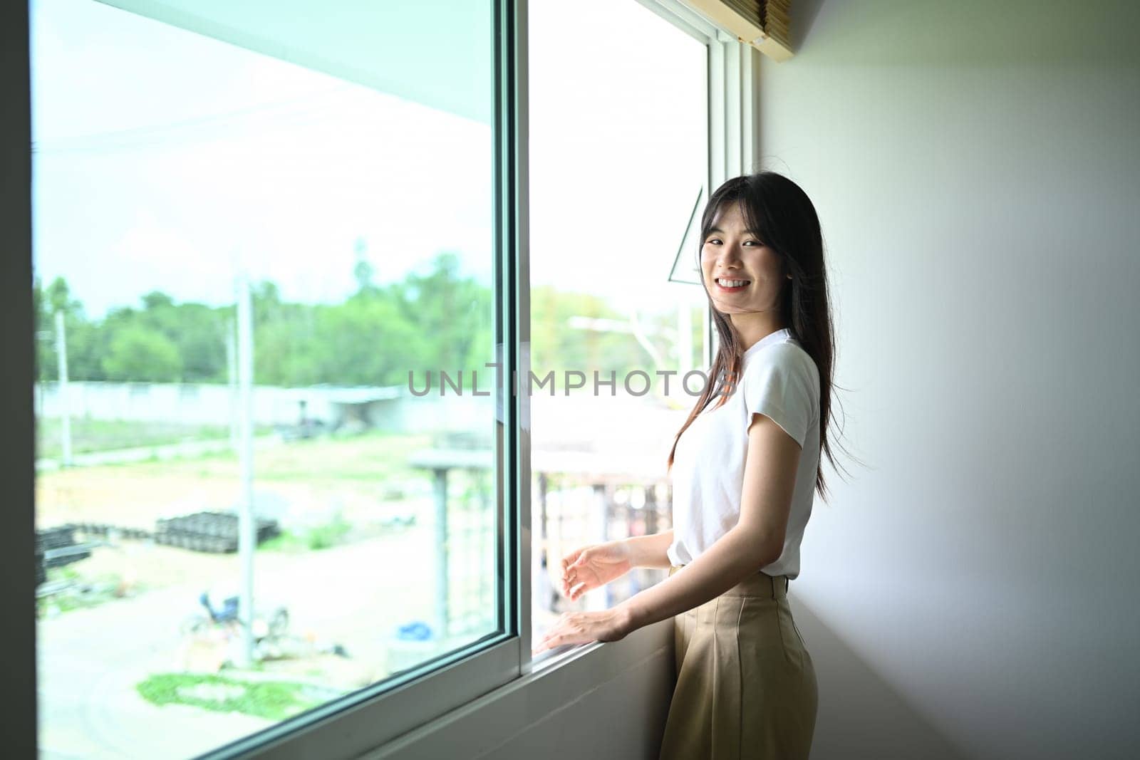 Cheerful young woman wearing casual clothes standing near window in bright room of her new house.
