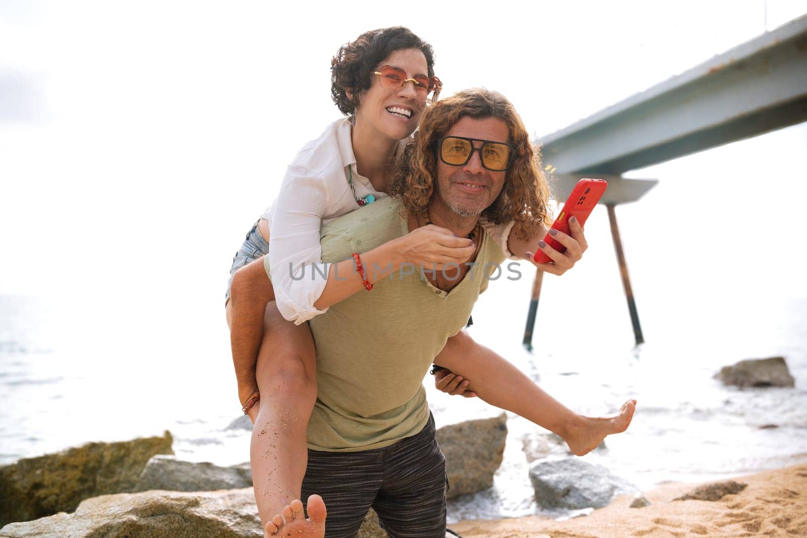 Happy caucasian man carrying his girlfriend on his back with smartphone, having fun together on the beach.