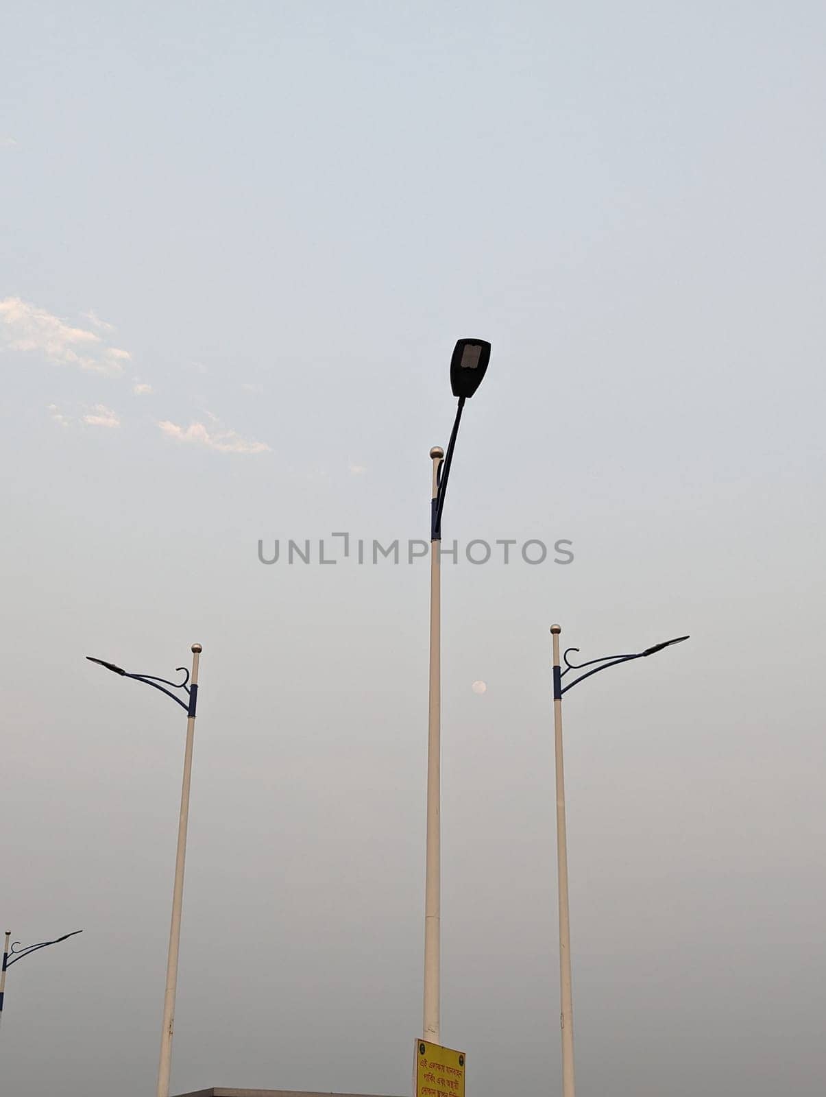 This image shows three streetlights against a hazy, pale blue sky at dusk. The streetlights are tall and slender, with white poles and black arms that extend out to the side. The streetlights are casting long shadows on the ground below. The sky is mostly clear, with just a few wisps of clouds visible. The overall mood of the image is peaceful and serene.