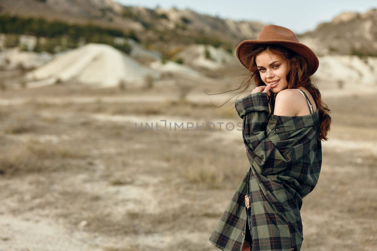 Woman in plaid shirt and hat standing in desert with majestic mountains in background on sunny day