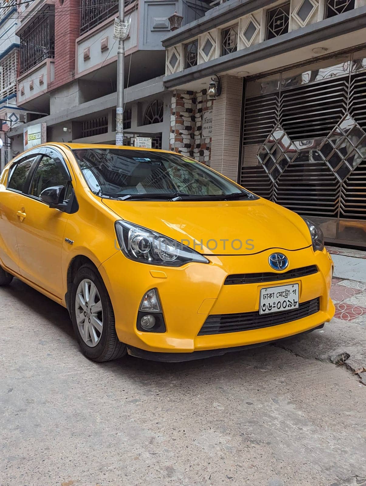 A yellow Toyota Prius C is parked on a street in Dhaka, Bangladesh. The car is facing forward and the front of the car is in focus. The car is parked on a street with a building in the background.