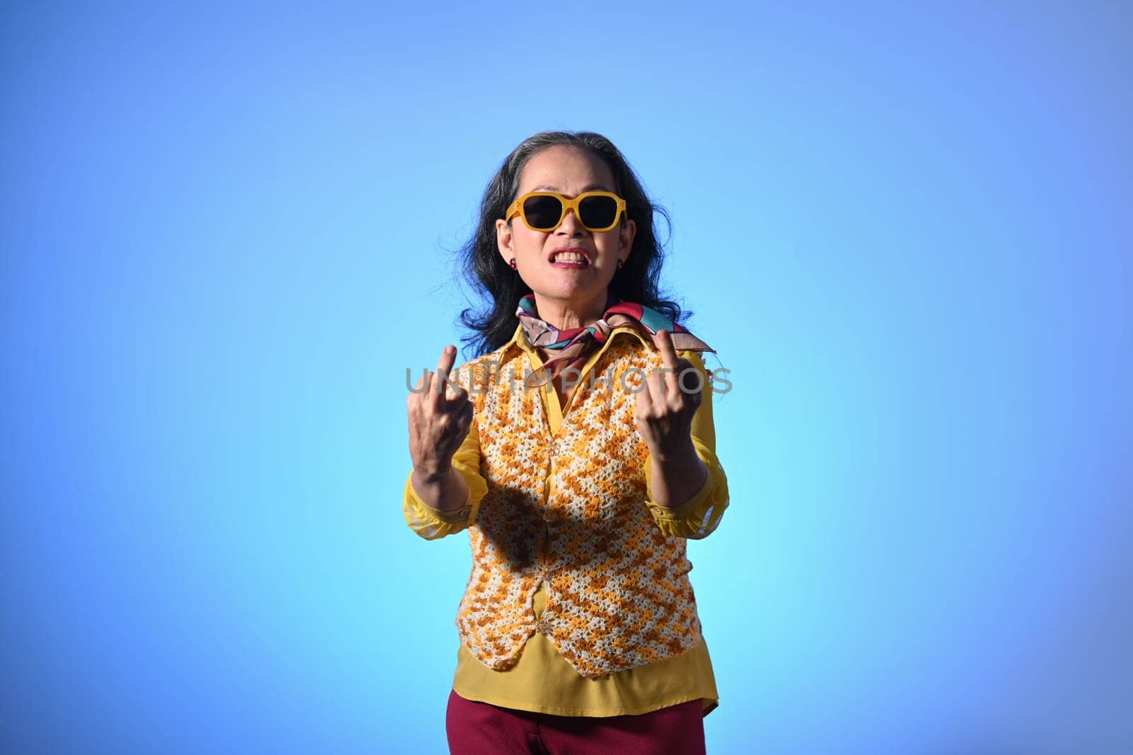 Crazy senior woman wearing sunglasses showing middle finger gesture over blue background.