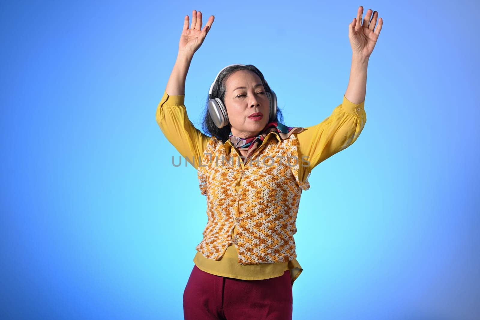 Joyful stylish middle age woman listening to music with headphones and dancing over blue background.