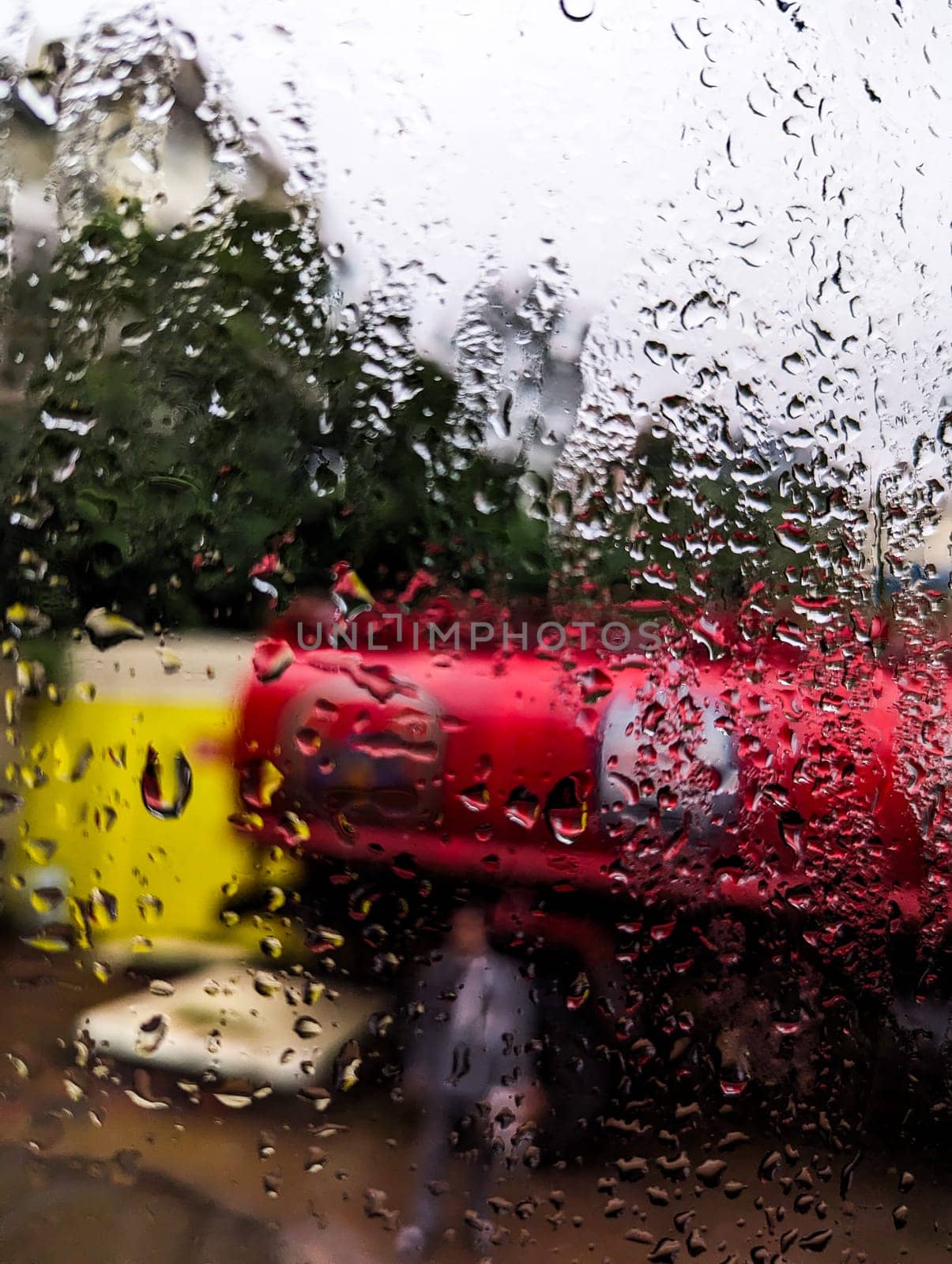 A blurry image of a red and yellow truck, likely a delivery vehicle, is seen through a window covered in rain droplets. The truck is parked in a city setting. The image captures the essence of a rainy day, with the raindrops blurring the background and creating a sense of movement. The image is taken from a perspective inside a vehicle or building.