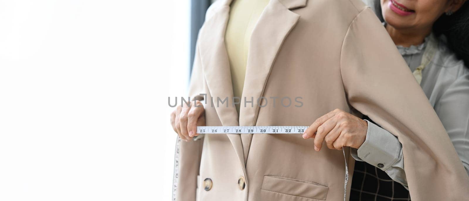 Skilled senior tailor working in the workshop studio measuring a jacket on a mannequin with tape measure.