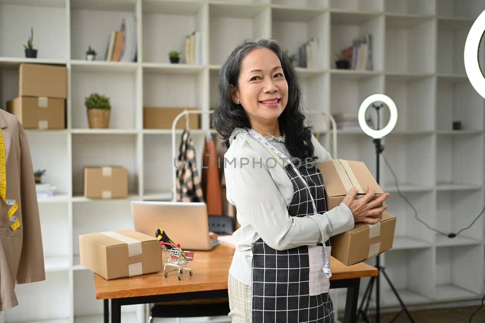 Confident senior woman small business owner holding parcel boxes of product and smiling to camera.