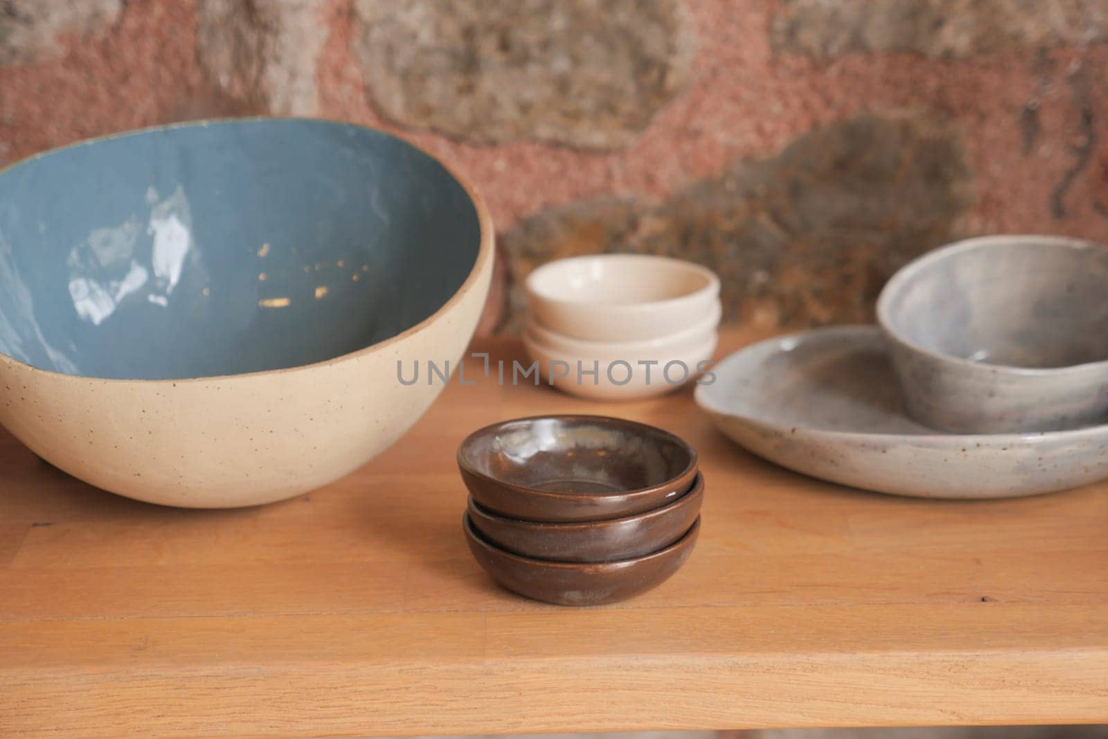 Rustic ceramic bowls and plates arranged on a wooden shelf, set against a stone wall in a kitchen or home interior