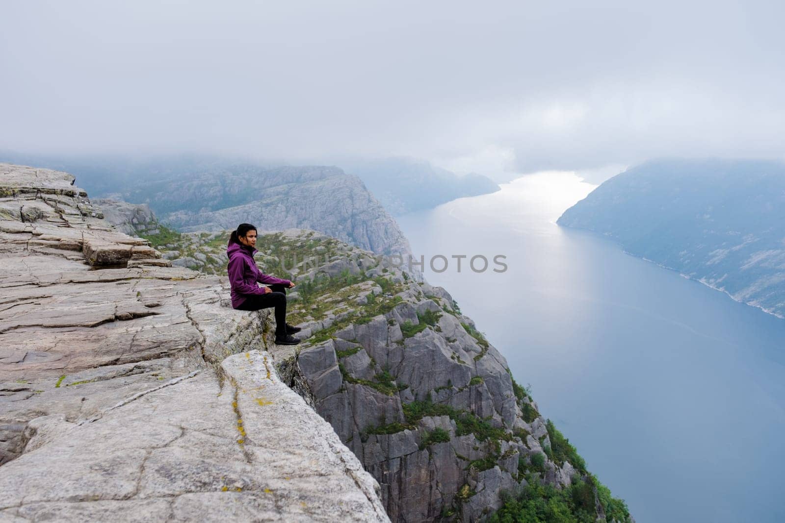 A Asian woman sits on the edge of the Preikestolen cliff in Norway, overlooking a stunning fjord. The misty morning adds to the dramatic landscape.