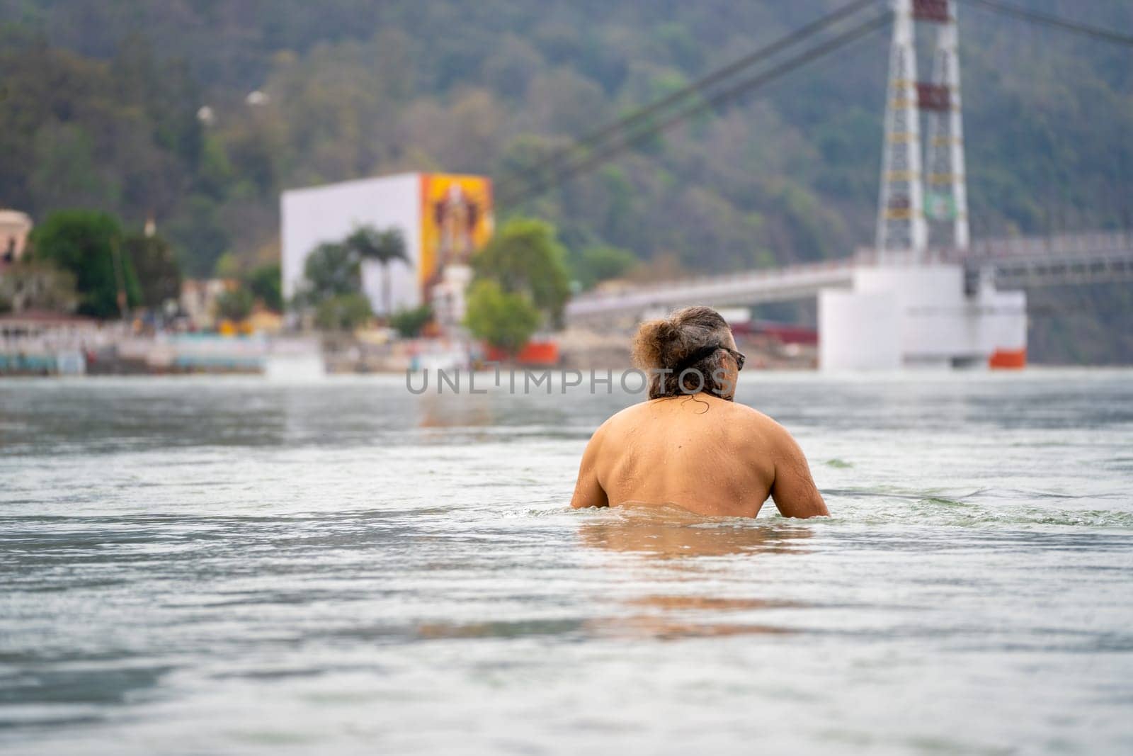 Haridwar, India - 30th Apr 2023: european man taking a dip in the river ganga with the ram jhula bridge in the distance showing the global appeal of this sacred town in India