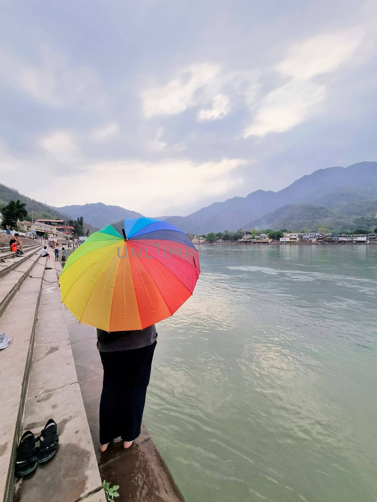 Indian woman standing on the banks of the river ganga on steps with a colorful umbrella and the himalaya mountains in distance in haridwar by Shalinimathur