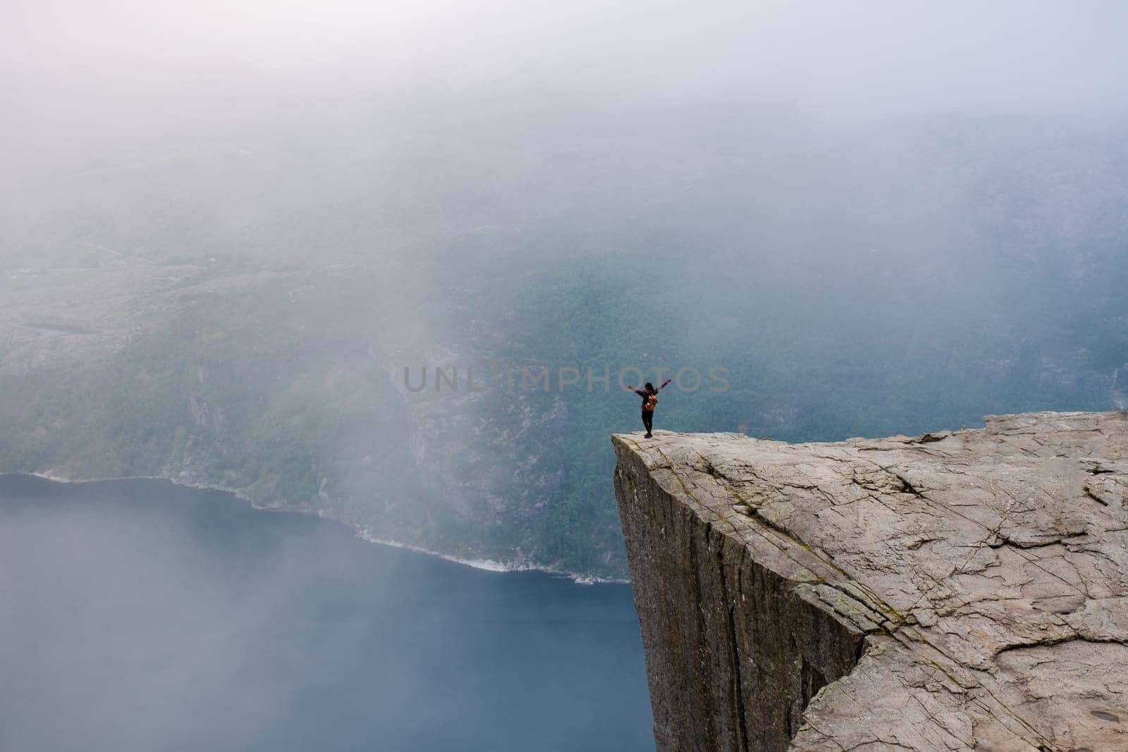 Preikestolen, Norway, A hiker stands on the edge of a cliff with arms outstretched, taking in the breathtaking view of the Norwegian fjords shrouded in mist by fokkebok