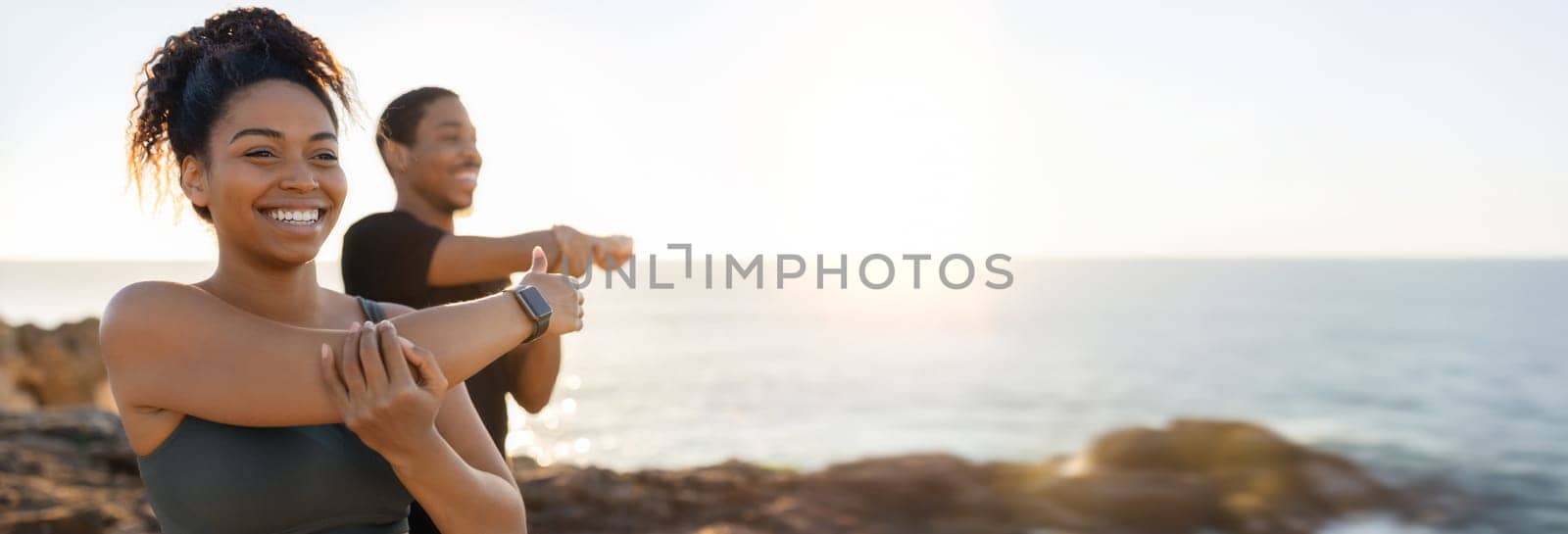 African American woman and man are stretching their arms in front of a beautiful sunset view over the ocean. They are standing on a rocky cliff, with the ocean stretching out behind them, copy space