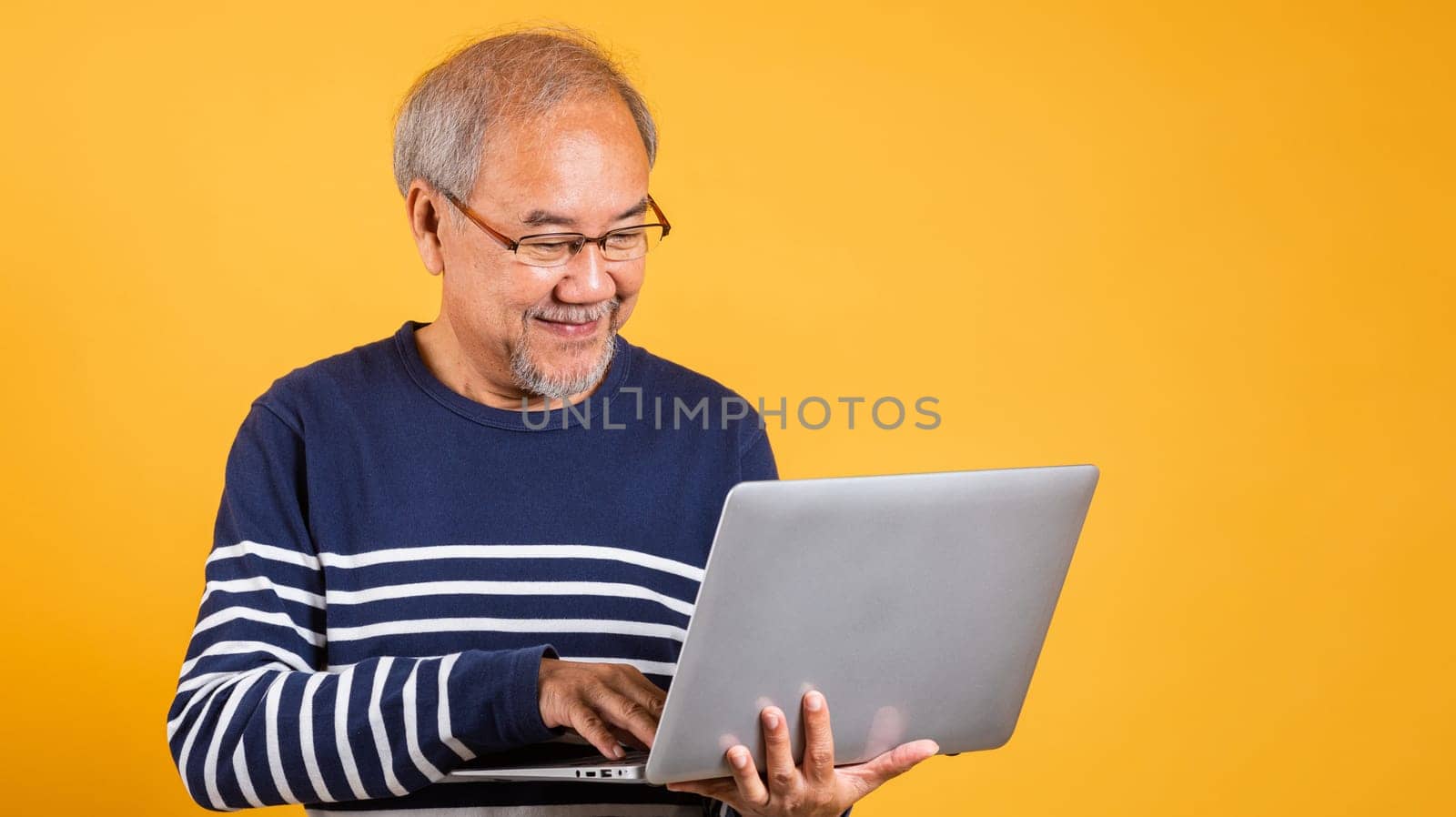 Portrait Asian old man wearing glasses use laptop computer studio shot isolated yellow background, smiling happy elderly man gray haired working, people lifestyle