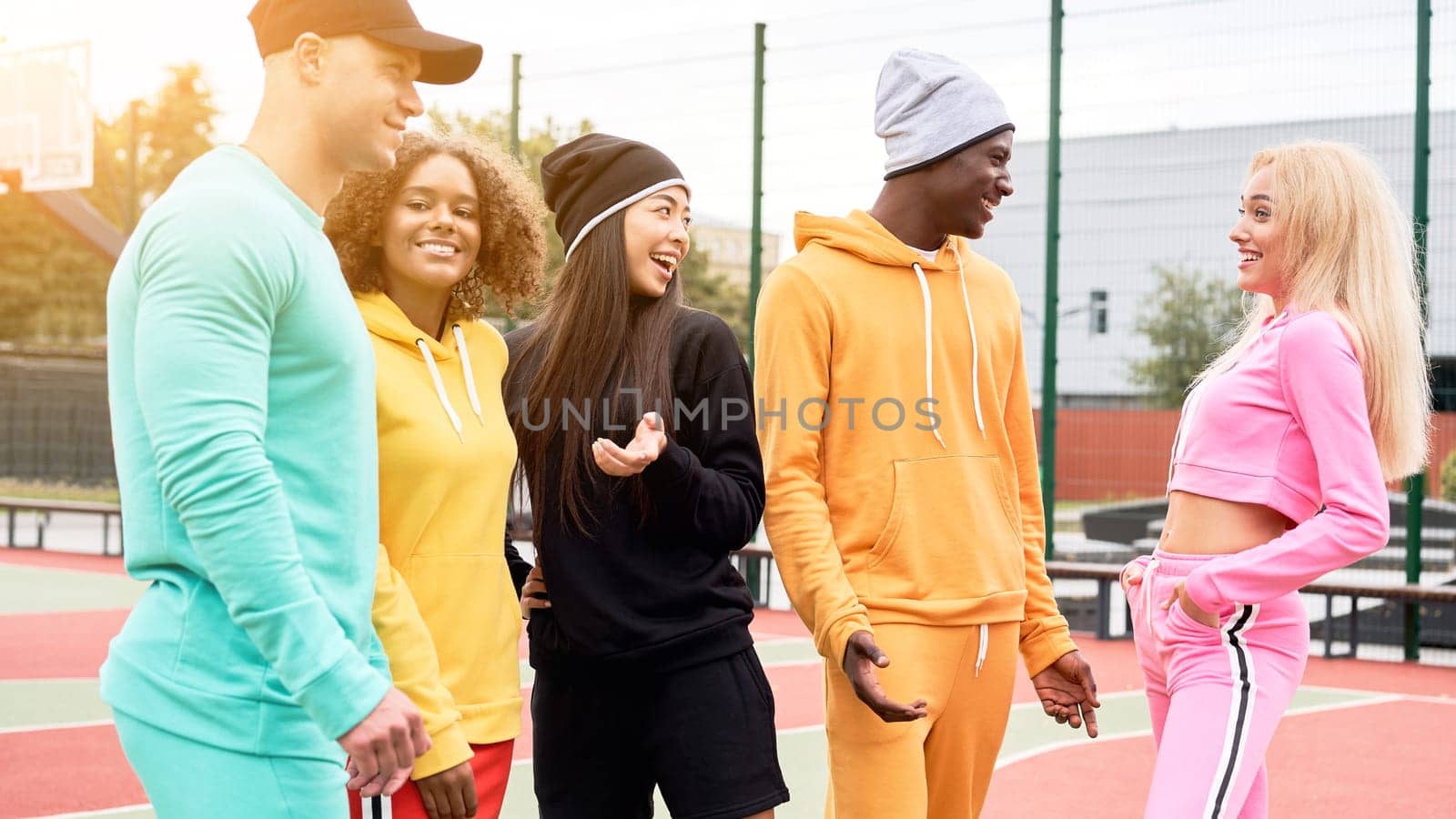 Group of young sportspeople of different nationalities in colorful sportswear talking at university stadium. Multi-ethnic group teenage friends spending time together at sportsground.