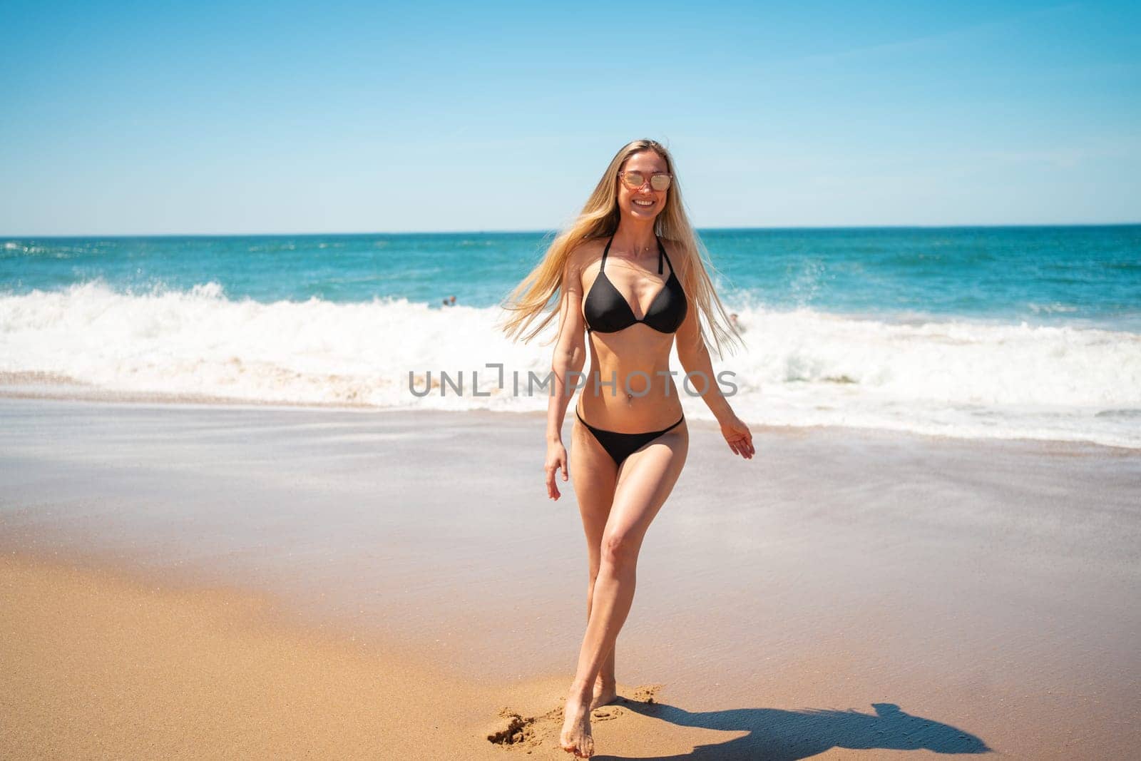 Full body of smiling young woman in bikini with long blond hair walking on sandy beach along sea waves. Attractive girl in swimwear near shoreline. Happy female is enjoying summer vacation.