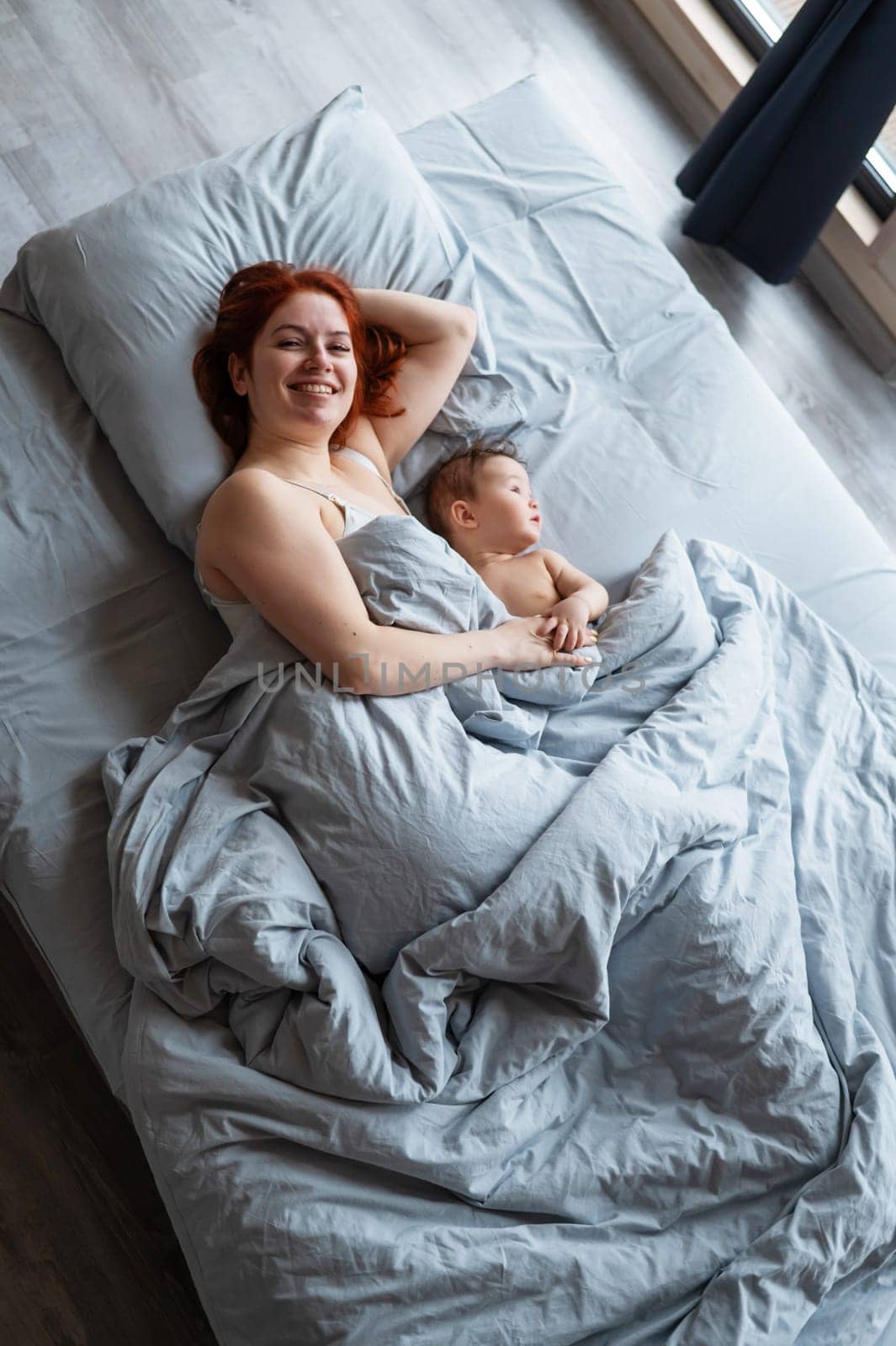 Top view of a red-haired Caucasian woman lying in bed with her baby son. Vertical photo