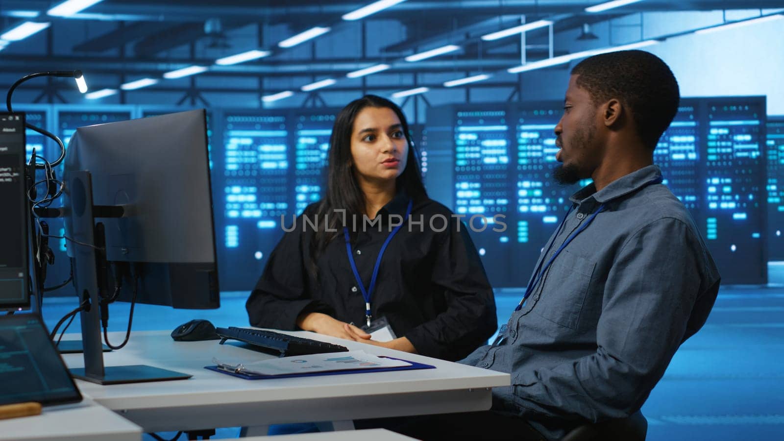 Multiracial team of IT engineers in server room brainstorming ways to fix equipment storing datasets. Indian and african american coworkers discussing how to mend racks doing computational operations