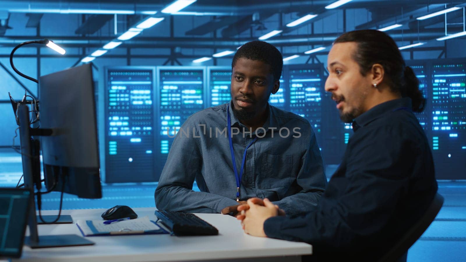 Multiracial engineers programming in high tech facility with server rows providing computing resources for different workloads. Diverse coworkers supervising data center supercomputers