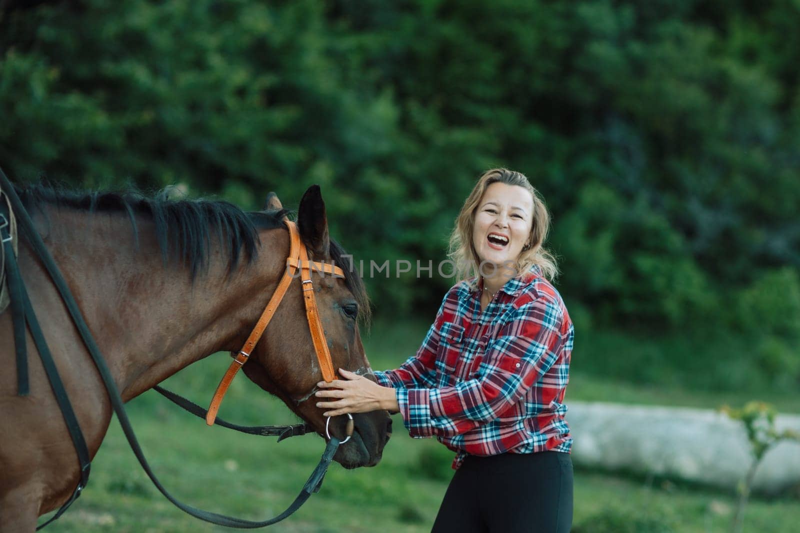 Happy blonde with horse in forest. Woman and a horse walking through the field during the day. Dressed in a plaid shirt and black leggings