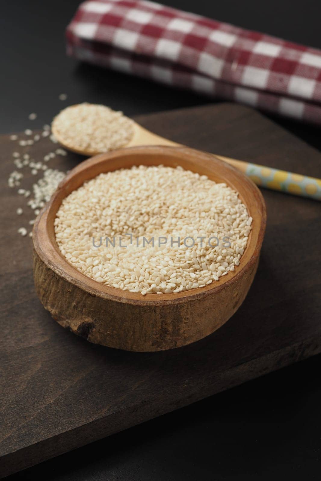 Close-up of white sesame seeds in a rustic wooden bowl and spoon, with a red plaid cloth in the background.