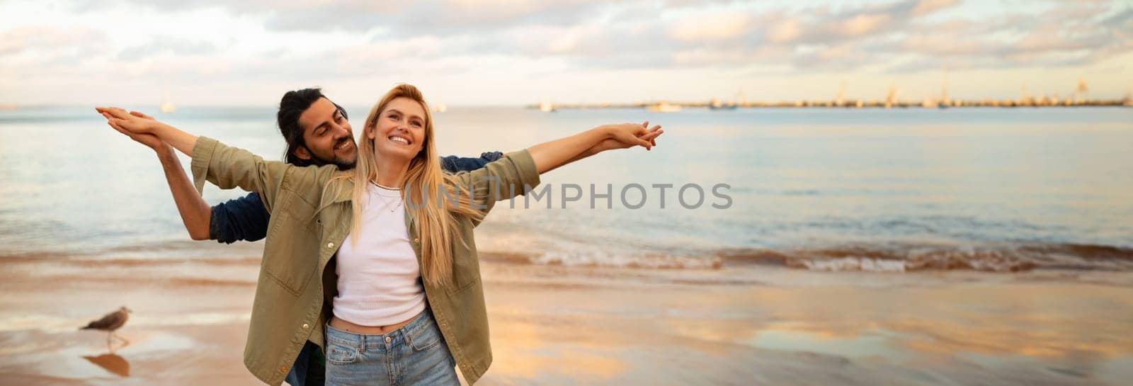 A couple enjoys the warmth of the evening sun as they stand together on a sandy beach, the woman with her arms outstretched and the man holding her from behind, panorama with copy space