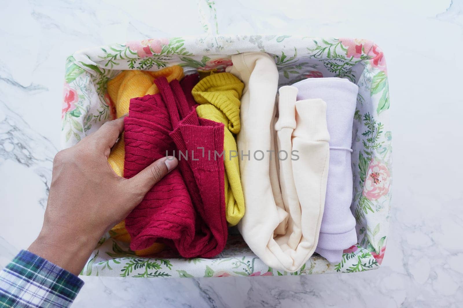 A hand arranging colorful clothes in a floral storage box on a marble surface, emphasizing organization and decor