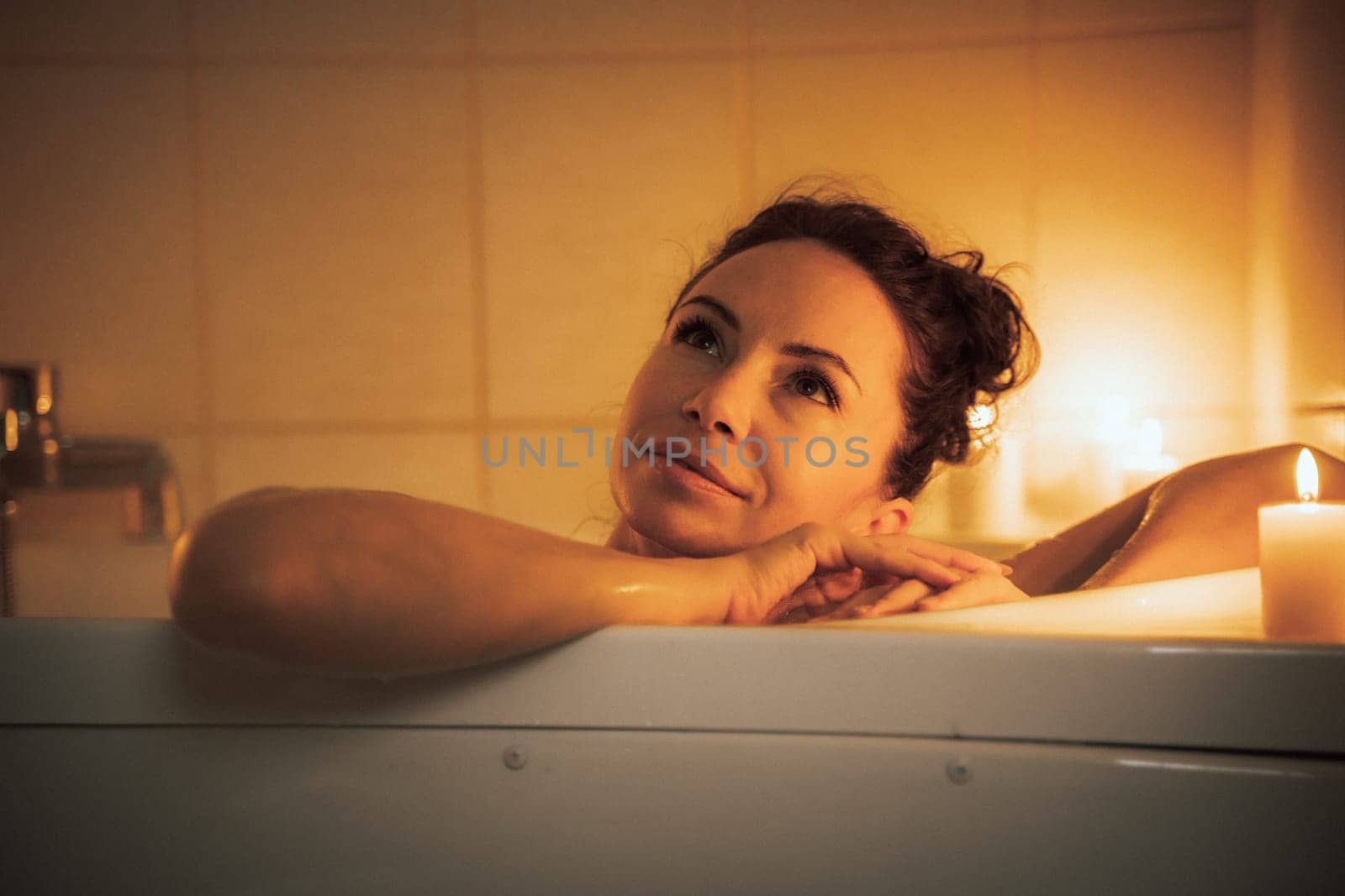 A woman is sitting in a bathtub with candles lit around her. She is smiling and she is enjoying her time