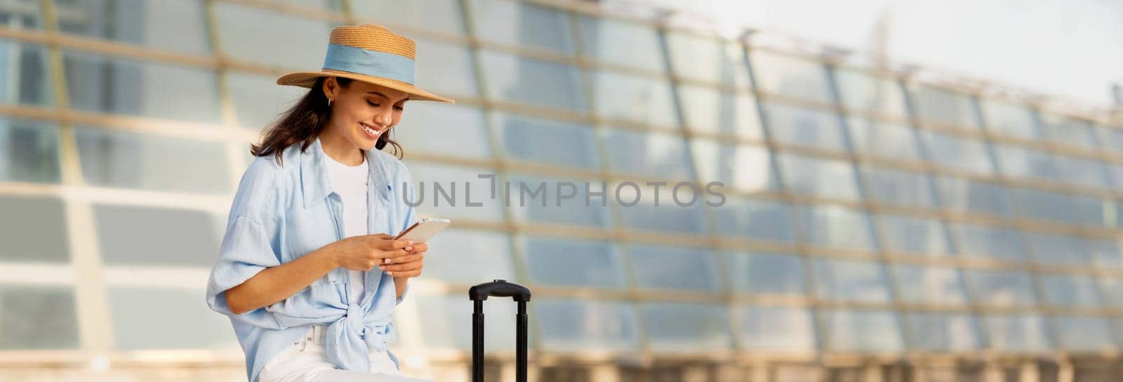 Smiling young european woman in hat with suitcase, typing on smartphone, enjoy trip at station, outdoor. App for tourist, travel with ticket, active lifestyle in summer with device, copy space