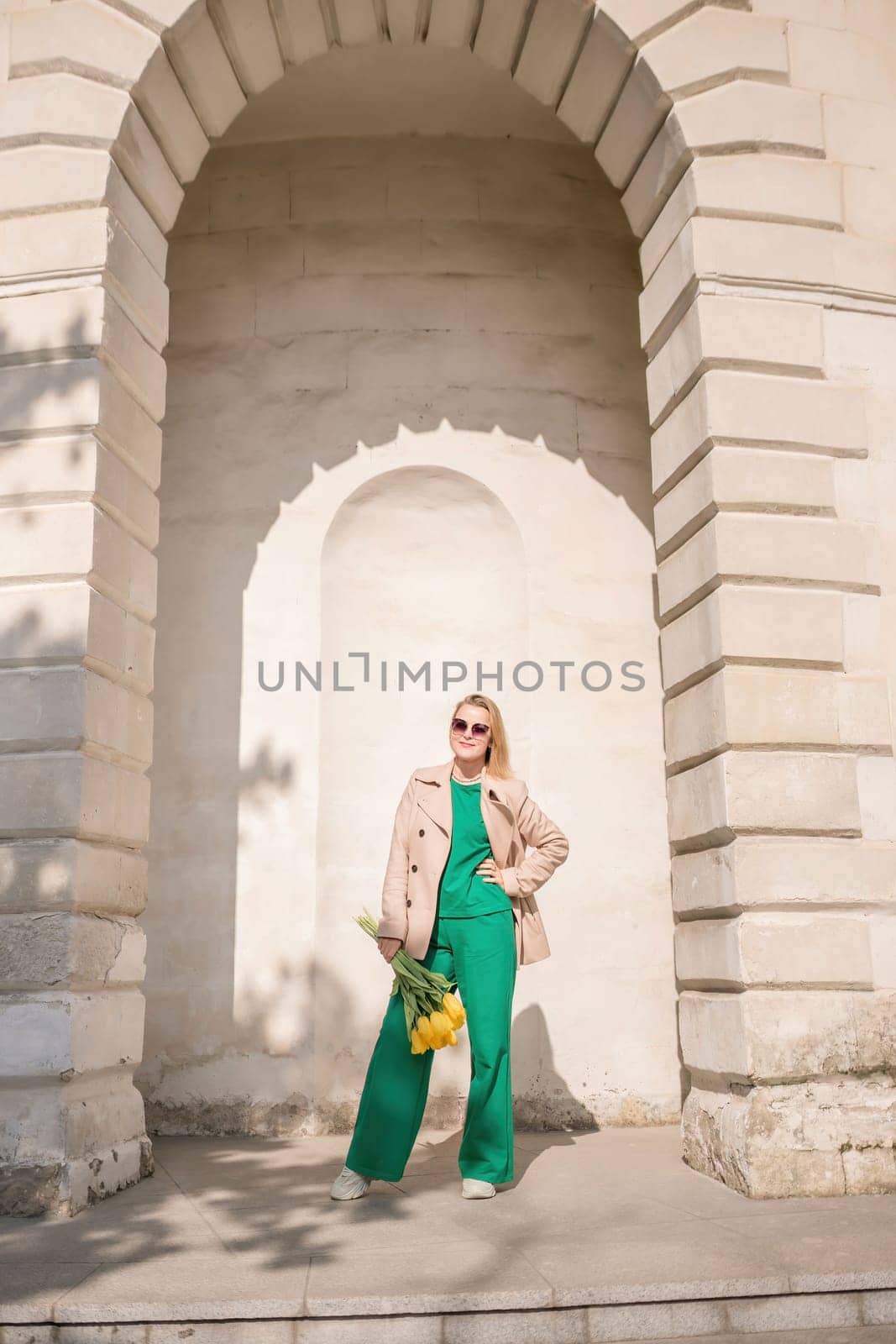 A woman is standing in front of a building with a yellow flower in her hand. She is wearing a green dress and a tan coat