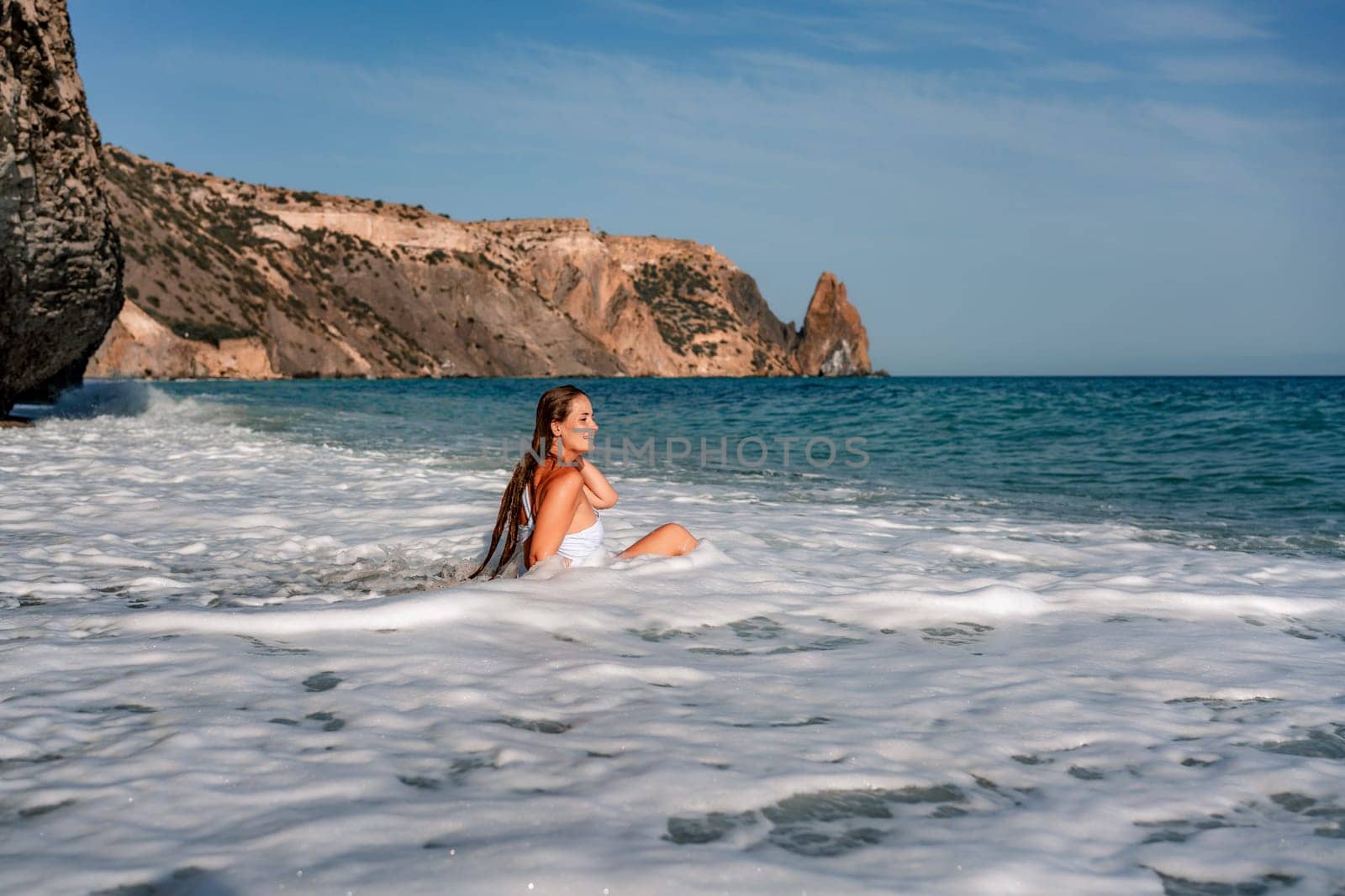 Happy woman in bikini sits on the sea beach. Tanned girl sunbathing on a beautiful shore. Summer vacation or holiday travel concept.