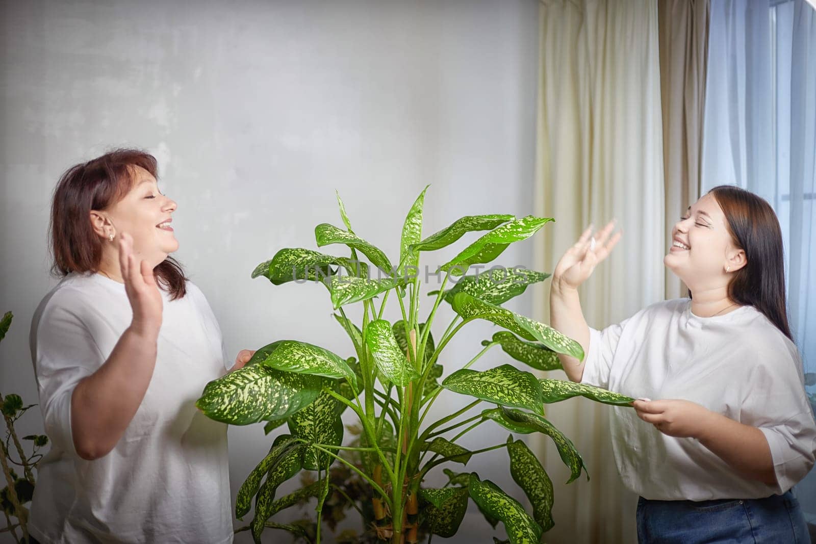 Happy Overweight family with mother and daughter in room with Diffenbachia. Middle aged woman and teenager girl having fun, joy by keleny