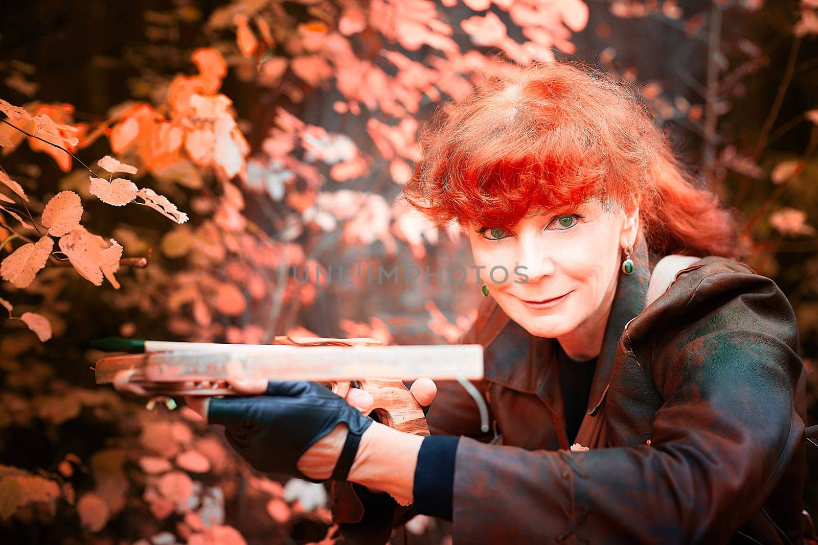 Mature model portraying a royal huntress with red curve hair is hunting with a crossbow in the in vibrant autumn forest in thematic photo shoot