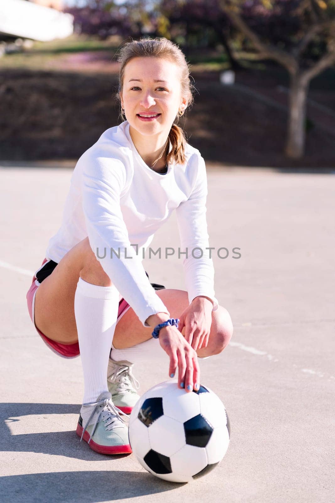 young caucasian woman squatting next to a soccer ball ready to play in a urban football court, concept of sport and active lifestyle