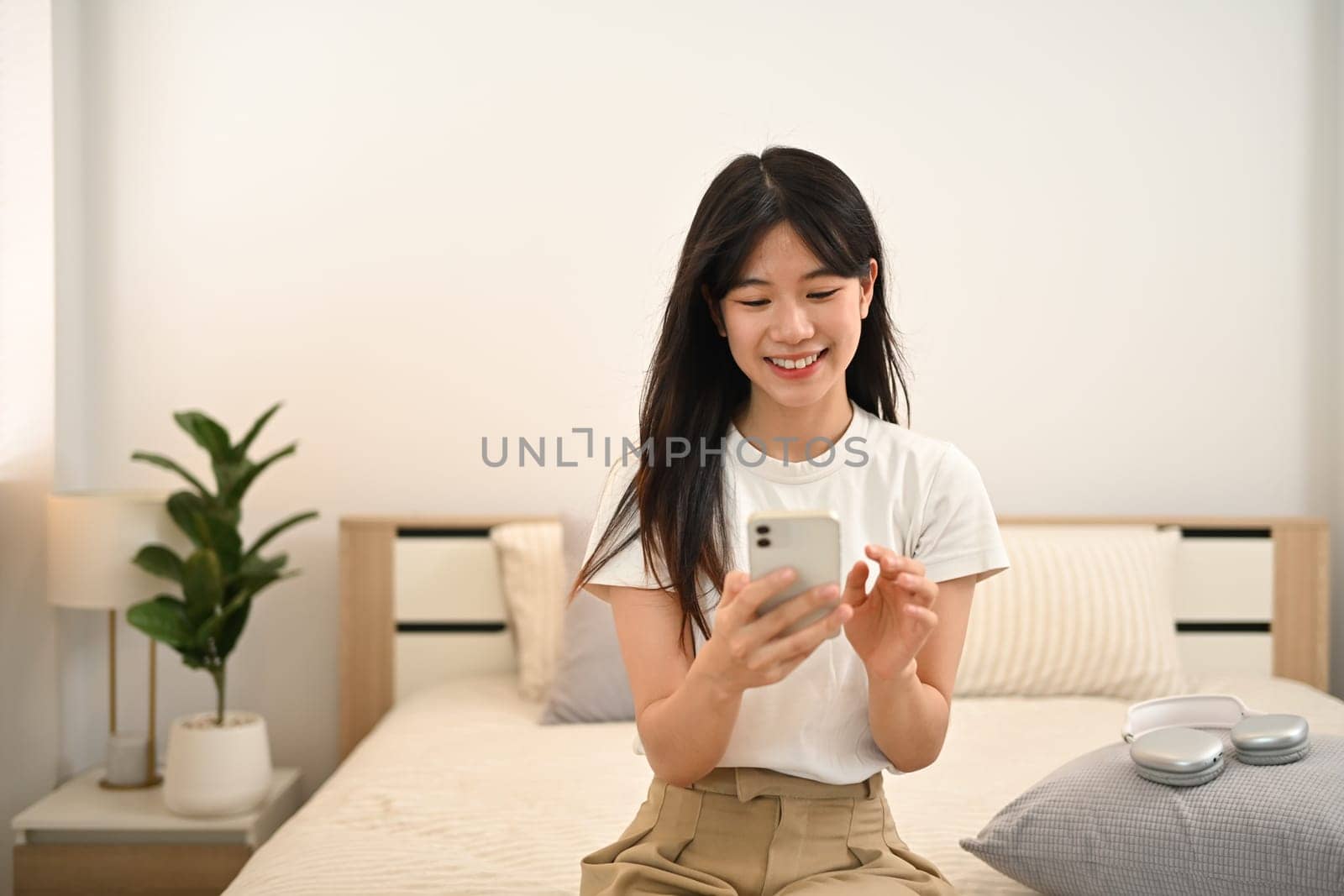 Smiling young woman using mobile phone sitting on bed at home.