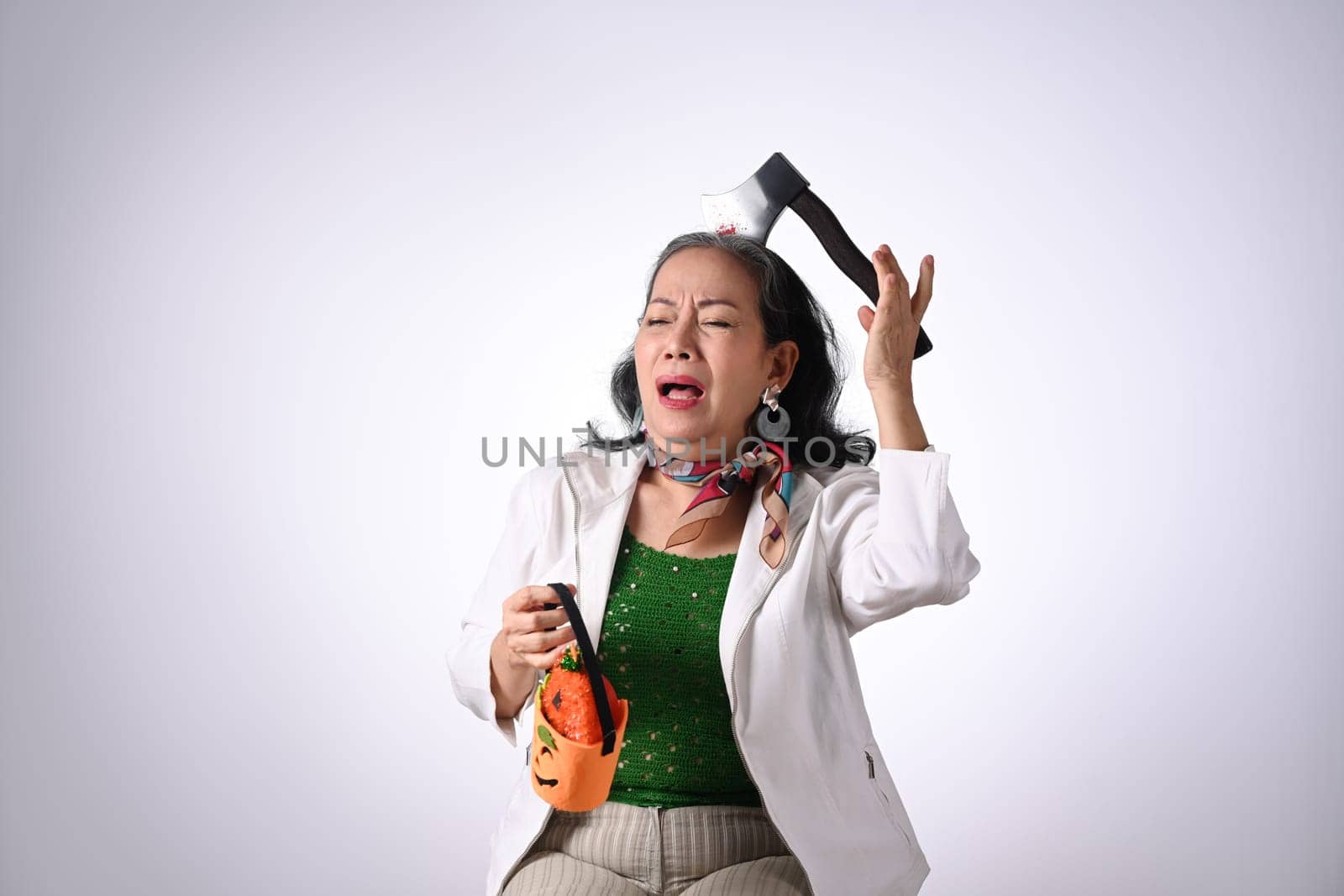 Playful senior woman dressed in halloween costume holding a pumpkin on white background.