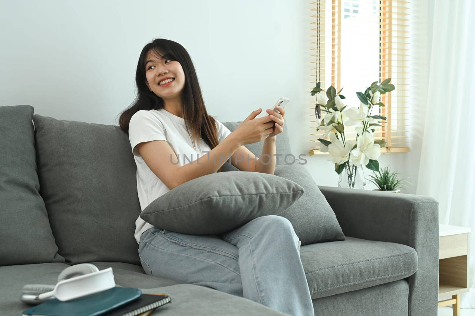 Relaxed young Asian woman using mobile phone on a couch at home by prathanchorruangsak