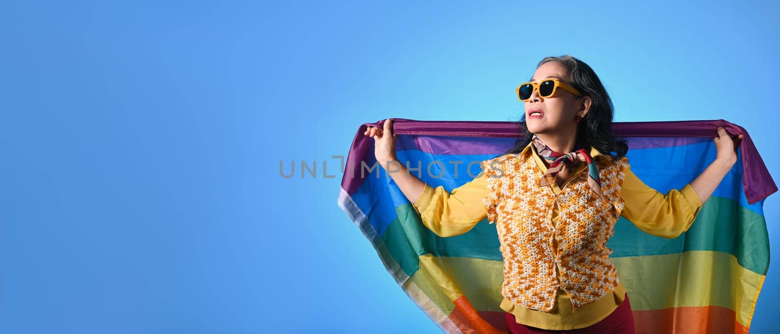 Elegant senior lady holding rainbow flag on blue background. LGBT, human rights, freedom and equality concept.