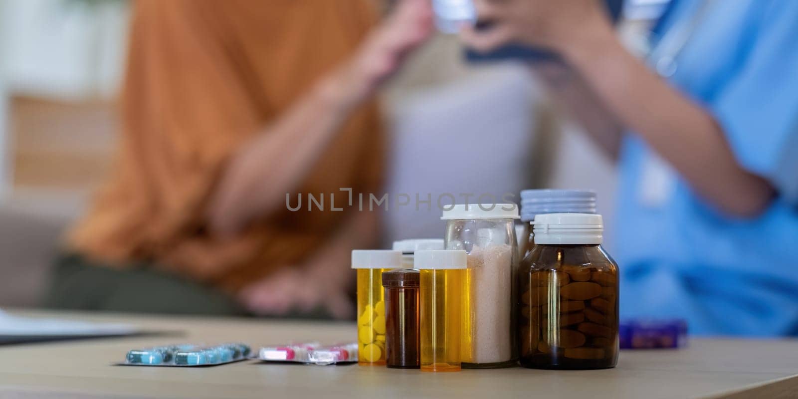 Close-up of various medication bottles and pills on a table with an elderly woman and caregiver in the background, highlighting elderly care and medication management