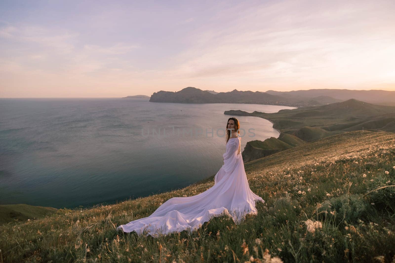 A woman in a white dress stands on a grassy hill overlooking the ocean. The scene is serene and peaceful, with the woman's dress flowing in the wind
