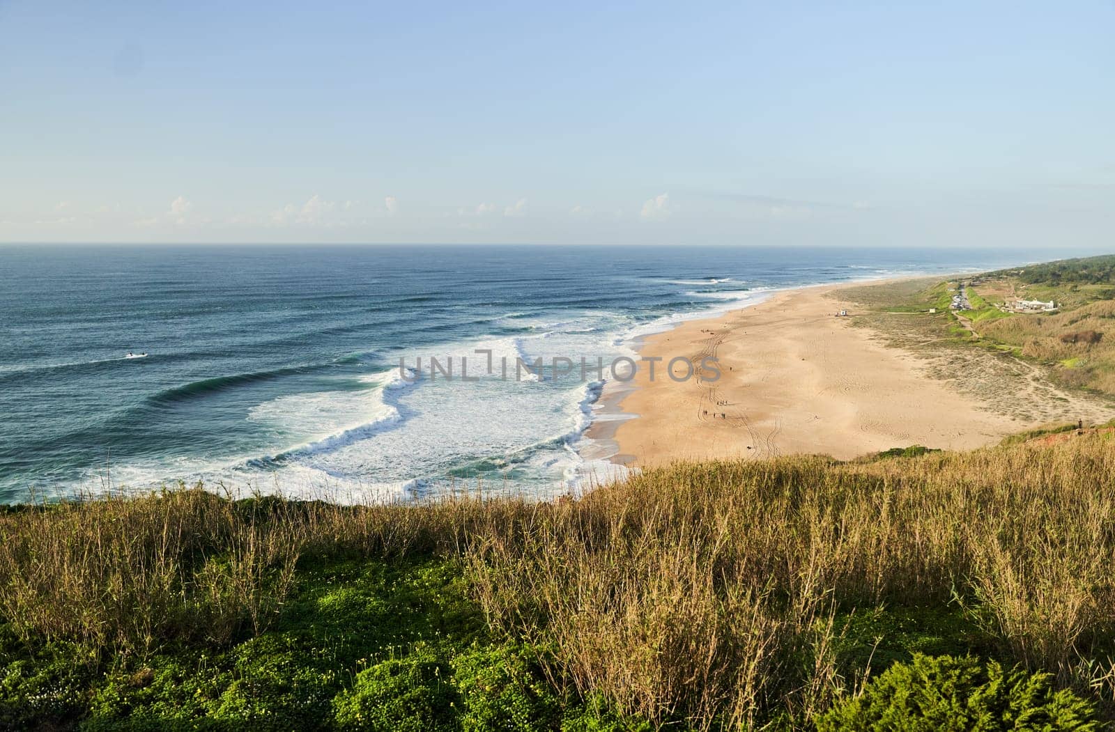 A breathtaking aerial view of a sandy beach and crystalclear ocean waters from a majestic cliff surrounded by lush greenery and coastal landforms. Nazare, Portugal