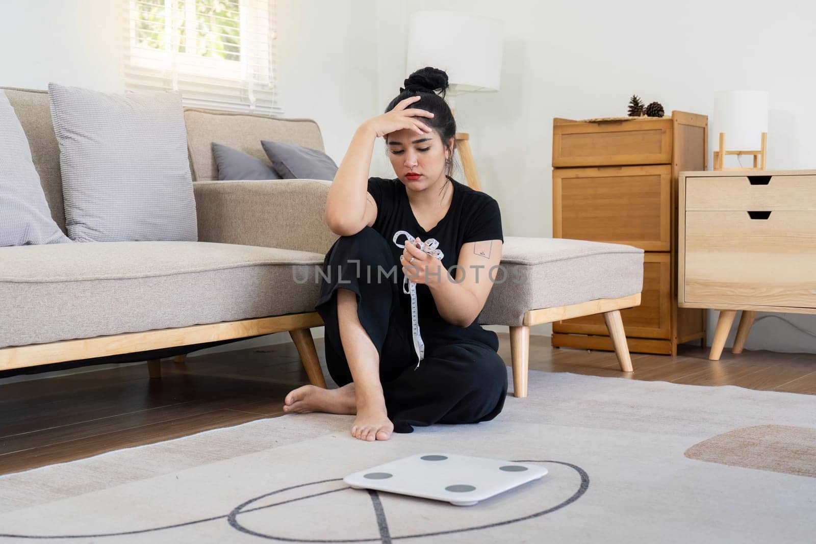 Stressed woman sitting on the floor holding a tape measure with a scale nearby, highlighting weight concern and anxiety